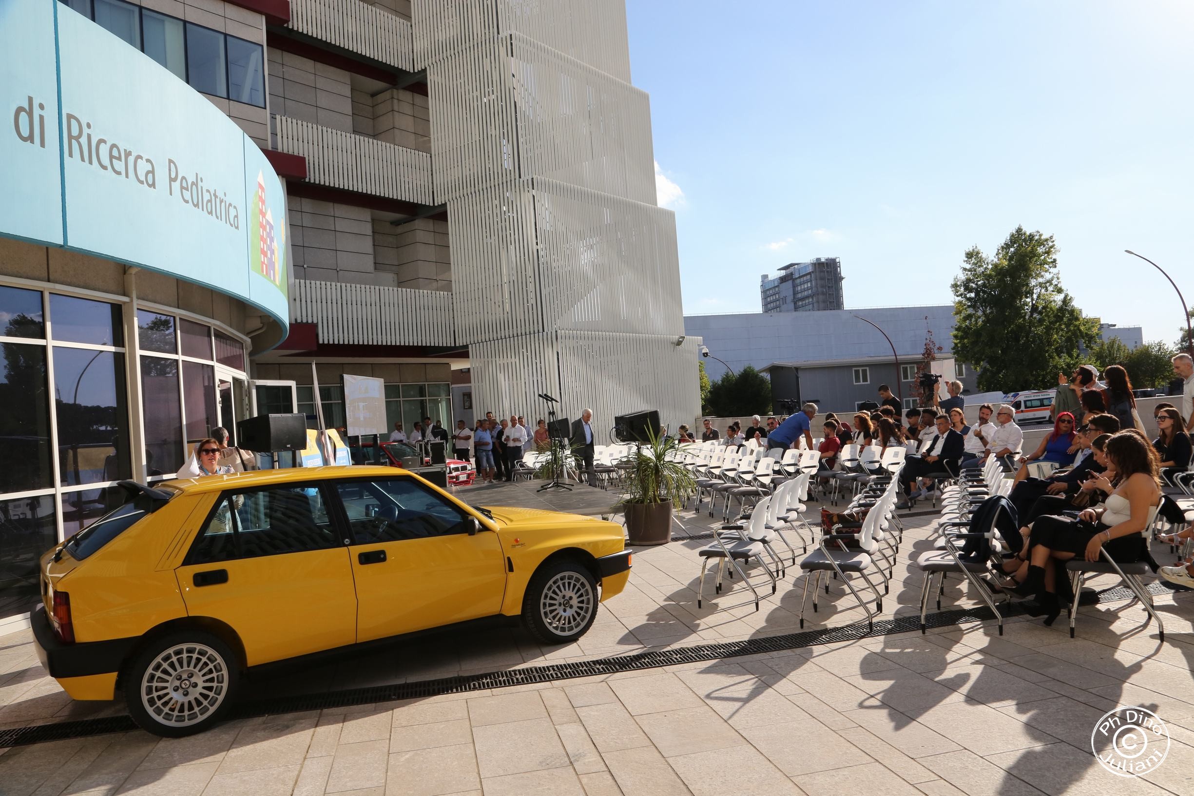 Preparativi per un evento speciale davanti alla Città della Speranza: tra il palco, le sedie bianche e il sole del pomeriggio, spicca una storica auto gialla