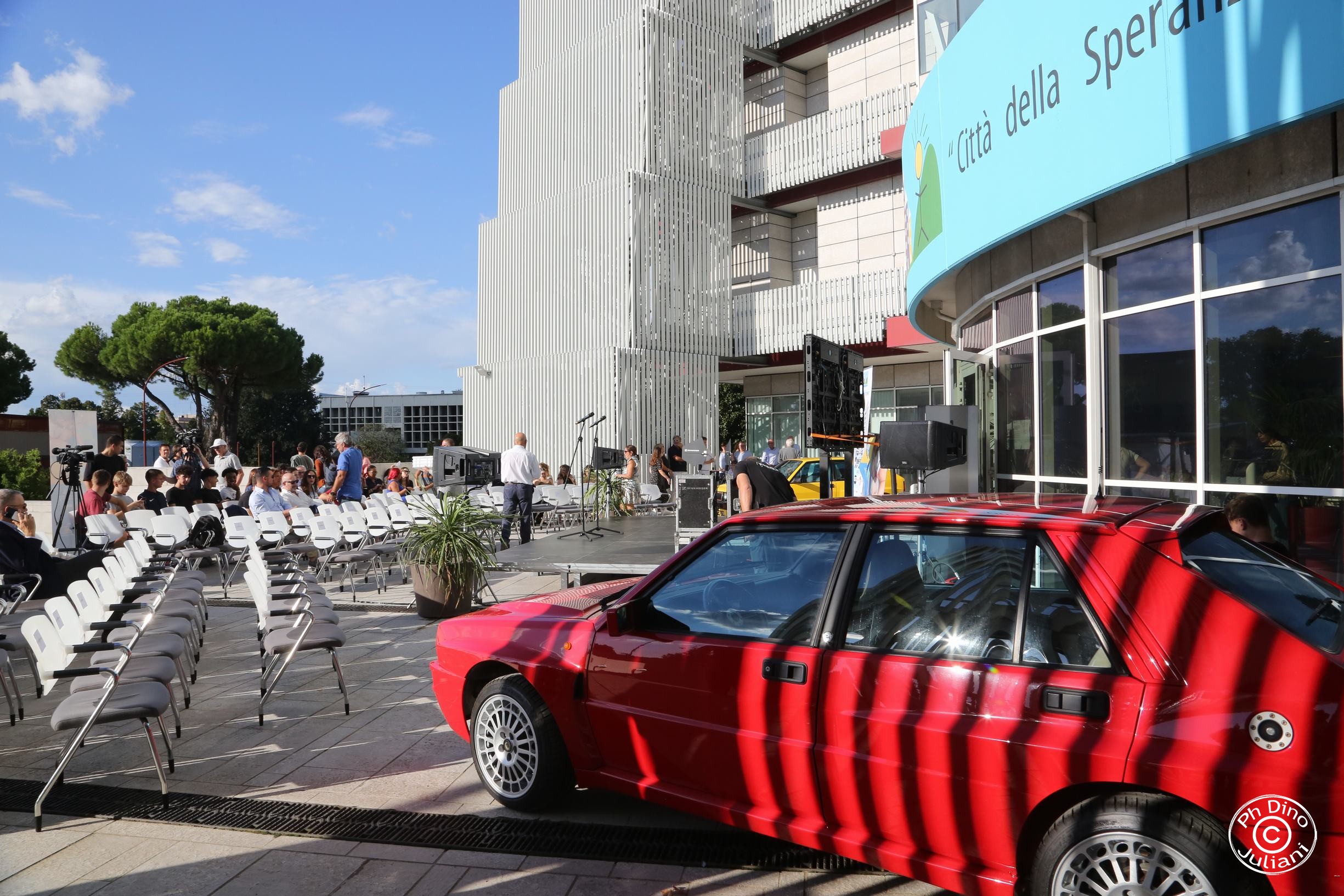 Allestimento di un evento all’aperto davanti alla sede della Città della Speranza, con palco, sedie e un’auto rossa parcheggiata in primo piano.