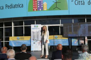 Presentatrice sorridente sul palco vestita di bianco davanti all'ingresso di Città della speranza