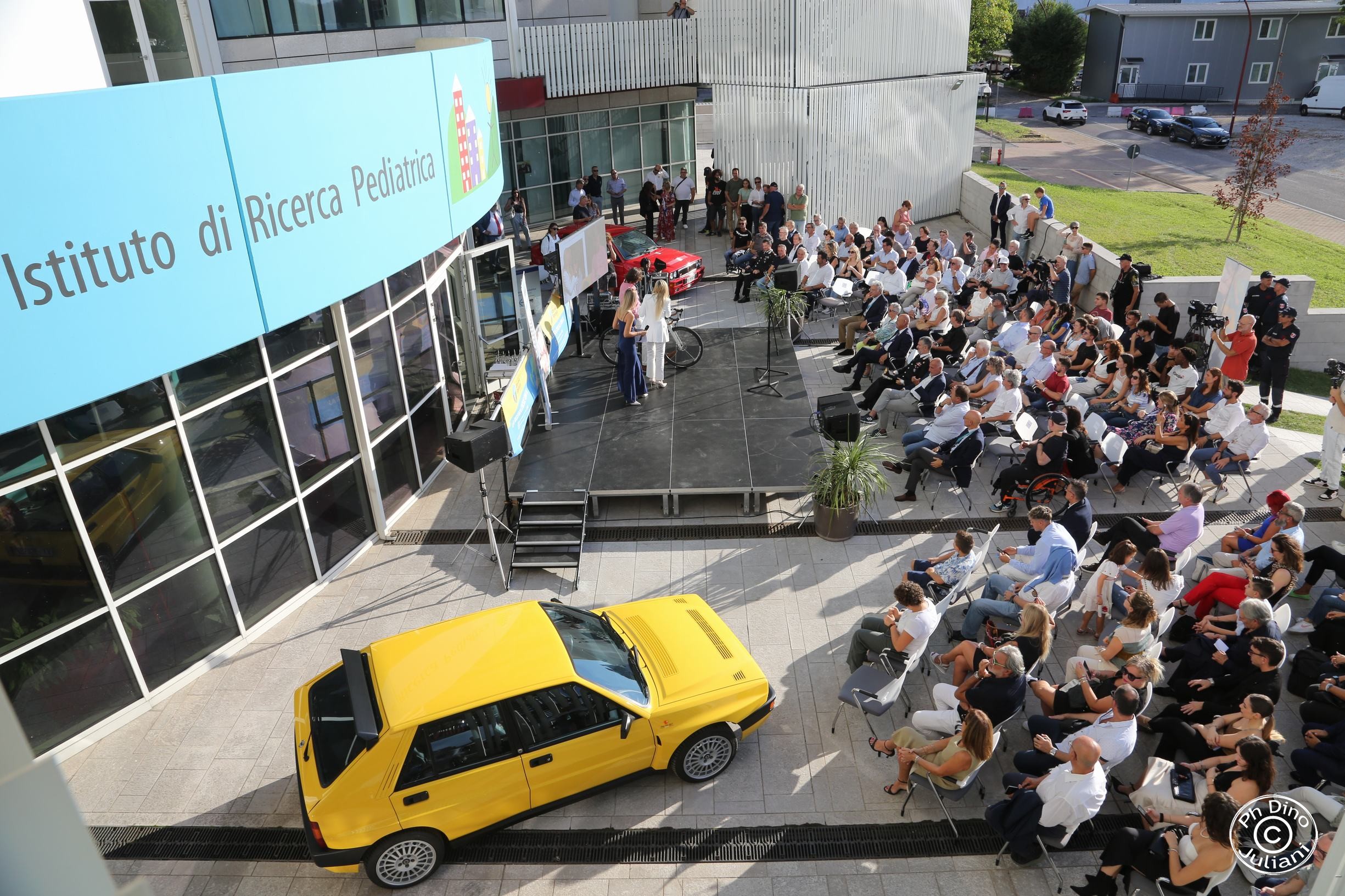 Evento all’esterno dell’Istituto di Ricerca Pediatrica, con palco, pubblico seduto e due auto d’epoca esposte: una gialla e una rossa.