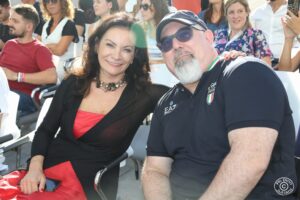 Due persone in prima fila per un evento istituzionale all’aperto. Dietro di loro, altre persone assistono all’evento.