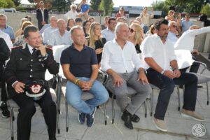 Diverse persone sedute in platea all’aperto durante un evento ufficiale. In prima fila, da sinistra a destra, un ufficiale dei Carabinieri in uniforme, un uomo con polo blu e jeans, due uomini in camicia bianca, e un altro uomo in camicia bianca con pantaloni scuri.