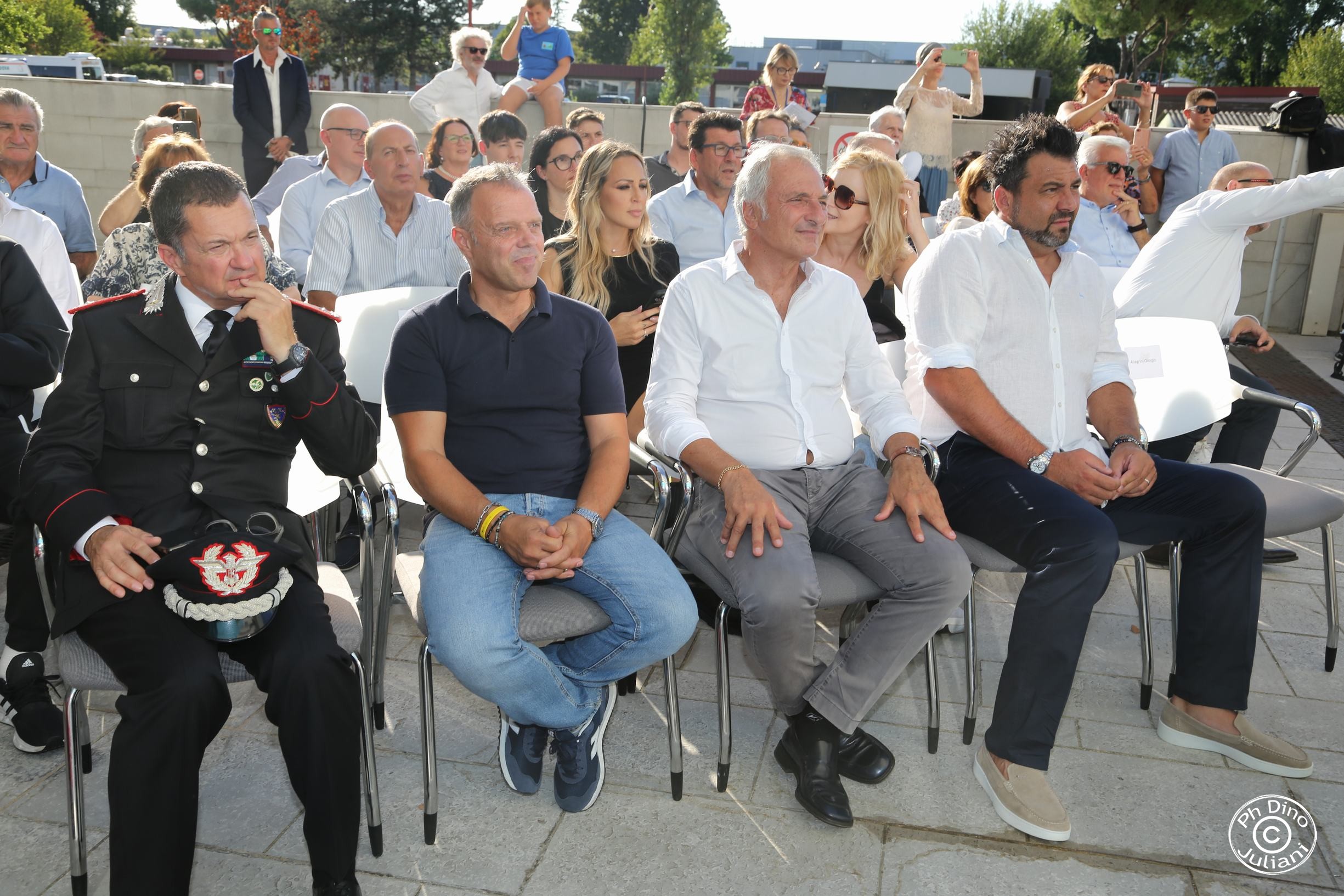 Diverse persone sedute in platea all’aperto durante un evento ufficiale. In prima fila, da sinistra a destra, un ufficiale dei Carabinieri in uniforme, un uomo con polo blu e jeans, due uomini in camicia bianca, e un altro uomo in camicia bianca con pantaloni scuri.