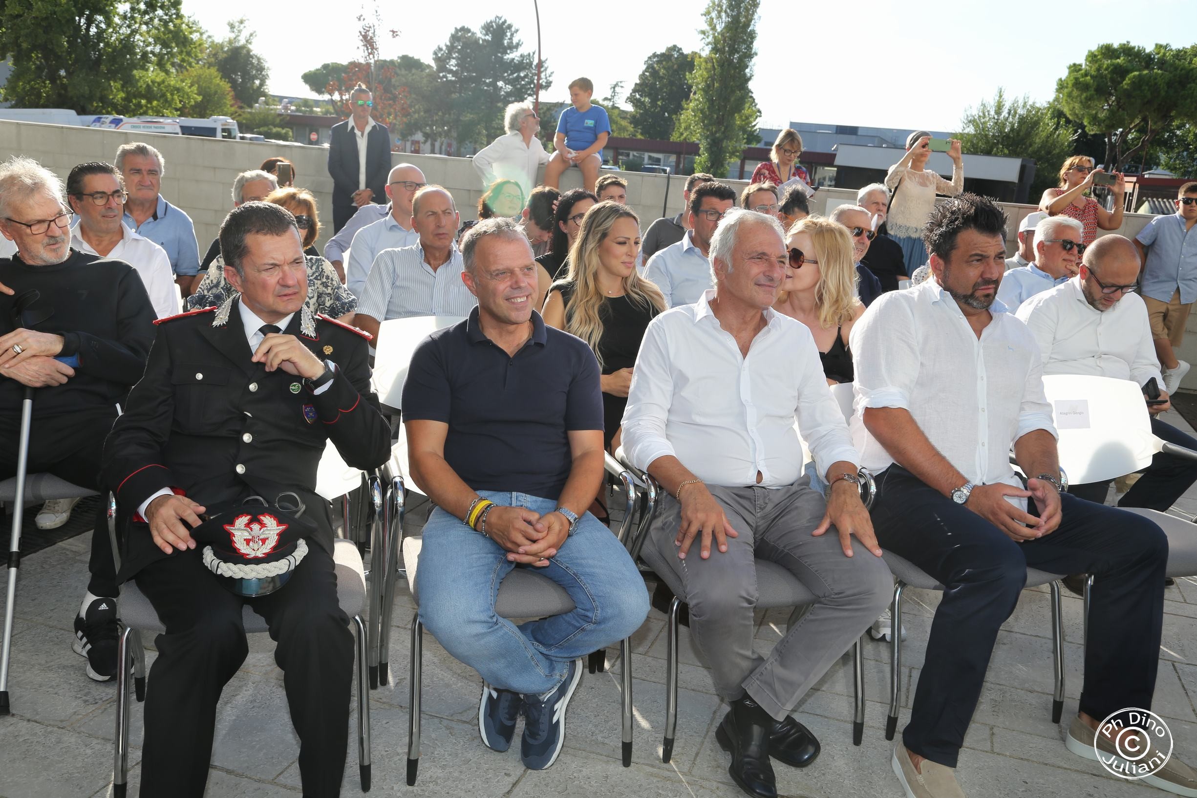 Pubblico seduto all’aperto durante un evento; in prima fila uomini in abbigliamento formale, tra cui un ufficiale dei Carabinieri.