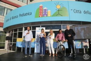 Cinque persone in piedi su un palco esterno durante un evento organizzato dalla Fondazione Città della Speranza.