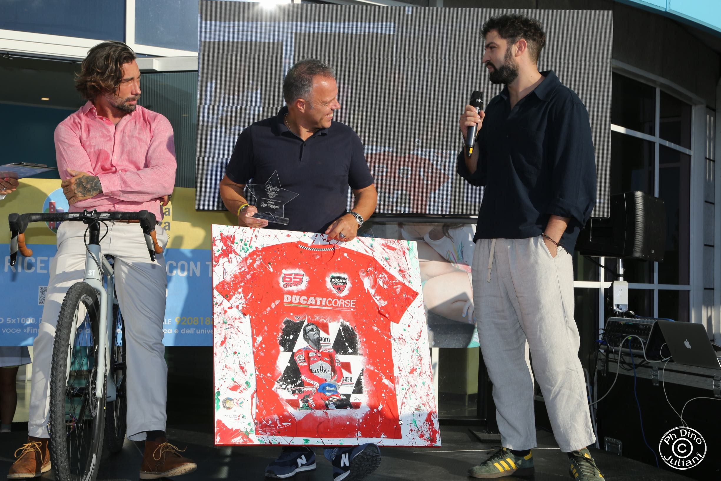 Un uomo sorridente in maglietta blu e jeans regge un trofeo trasparente a forma di stella con incisa una dedica. Con l'altra mano mostra un quadro raffigurante una maglietta rossa Ducati Corse con il numero 63 e un ritratto del pilota motociclistico in gara.