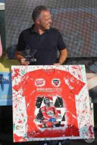 Emozionato e sorridente, un uomo riceve un premio speciale e mostra con orgoglio un’opera d’arte dedicata alla Ducati Corse