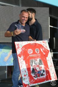 Uomo sorridente tiene un trofeo a forma di stella e un quadro con una maglietta rossa Ducati Corse, durante un evento pubblico.