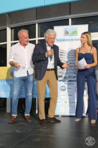 Un uomo in camicia bianca e jeans regge un trofeo trasparente a forma di stella, accanto a lui un altro uomo con capelli bianchi parla al microfono indossando giacca blu e pantaloni beige. A destra una giovane donna in abito blu tiene fogli in mano.