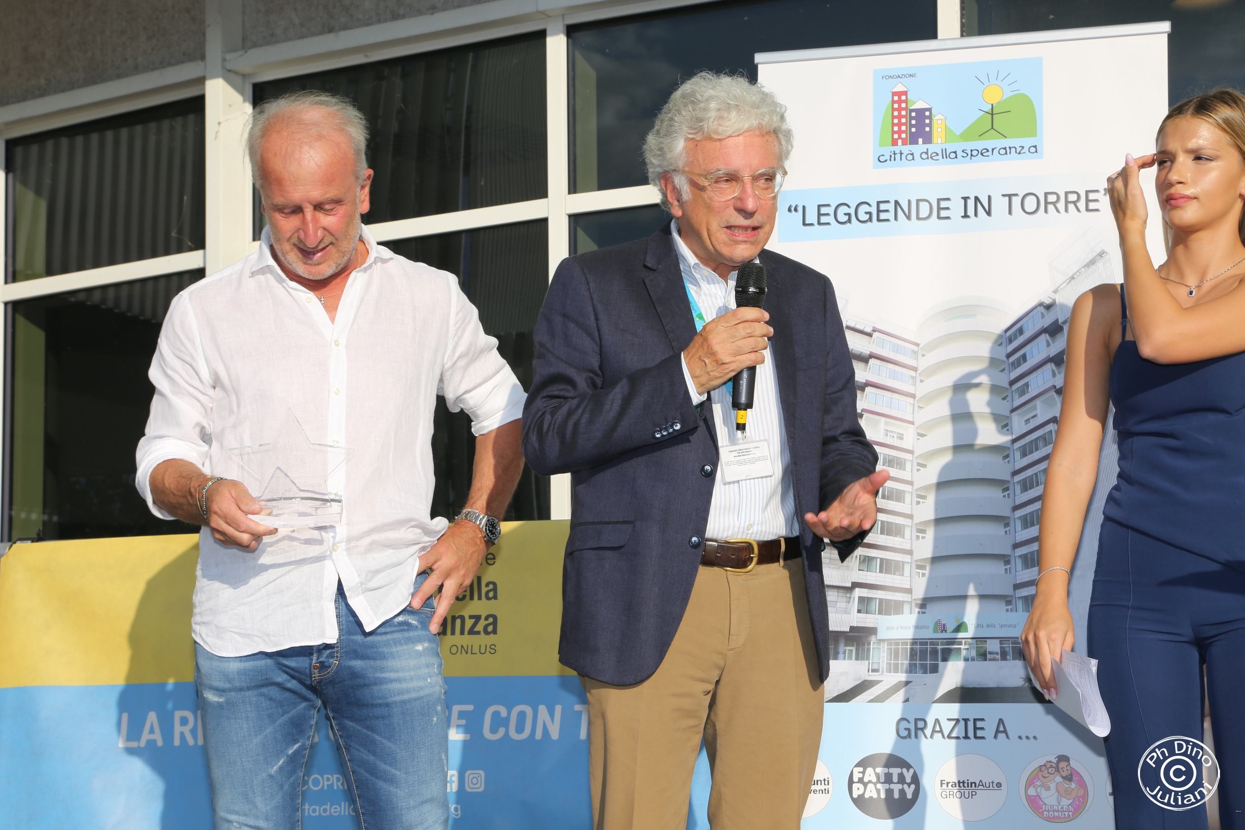 Tre persone sul palco durante un evento della Fondazione Città della Speranza. Un uomo a sinistra tiene un premio a forma di stella, l’uomo al centro parla al microfono, mentre la donna a destra ascolta con attenzione.