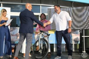 Sul palco della Fondazione “Città della Speranza”, un gruppo di ospiti prende parte a un evento pubblico. Una conduttrice vestita elegantemente, due uomini che si danno la mano e un uomo seduto su una bicicletta