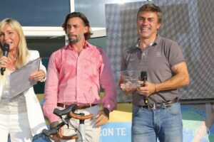 tre persone sul palco: una presentatrice con abito bianco, un uomo in camicia rosa che tiene un microfono e una bicicletta da corsa, e un altro uomo in polo grigia e jeans.