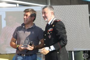 Cerimonia pubblica all’aperto con un ufficiale dei Carabinieri e un personaggio noto n un contesto legato a sport, beneficenza o sensibilizzazione sociale. Logo del fotografo visibile in basso a destra.