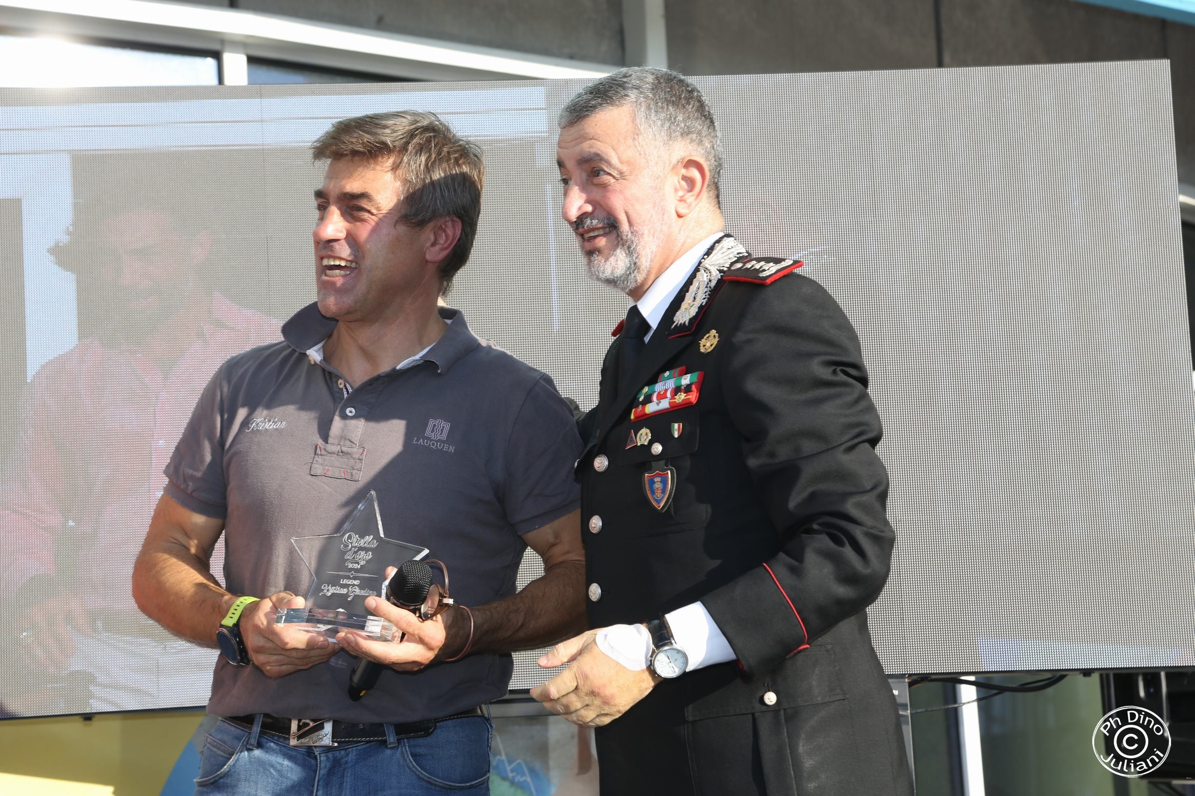 Cerimonia pubblica all’aperto con un ufficiale dei Carabinieri e un personaggio noto n un contesto legato a sport, beneficenza o sensibilizzazione sociale. Logo del fotografo visibile in basso a destra.