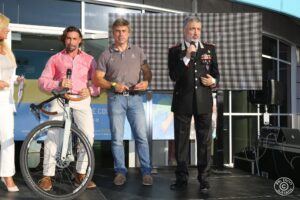 Quattro persone sul palco durante un evento pubblico: una donna in bianco, due uomini in abiti casual, e un ufficiale dei Carabinieri in uniforme che parla al microfono.