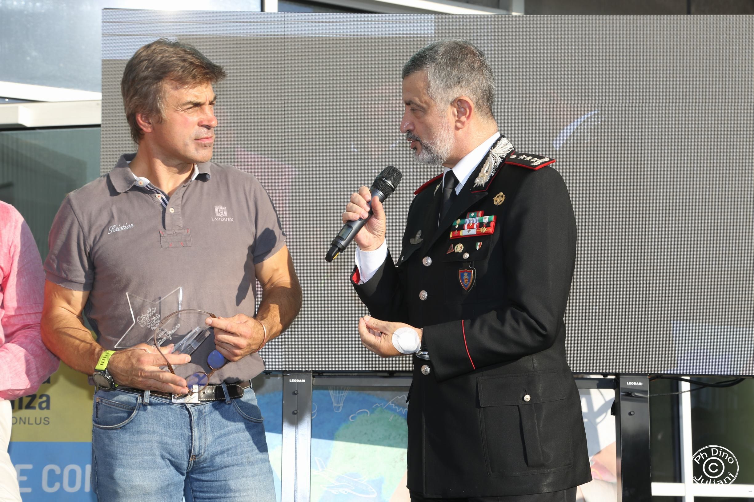 un uomo in uniforme sta parlando al microfono a un'altra persona che tiene in mano un premio