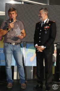 due persone sul palco, uno vestito in uniforme sorridente. L'altro tiene un premio a forma di stella e parla al microfono.