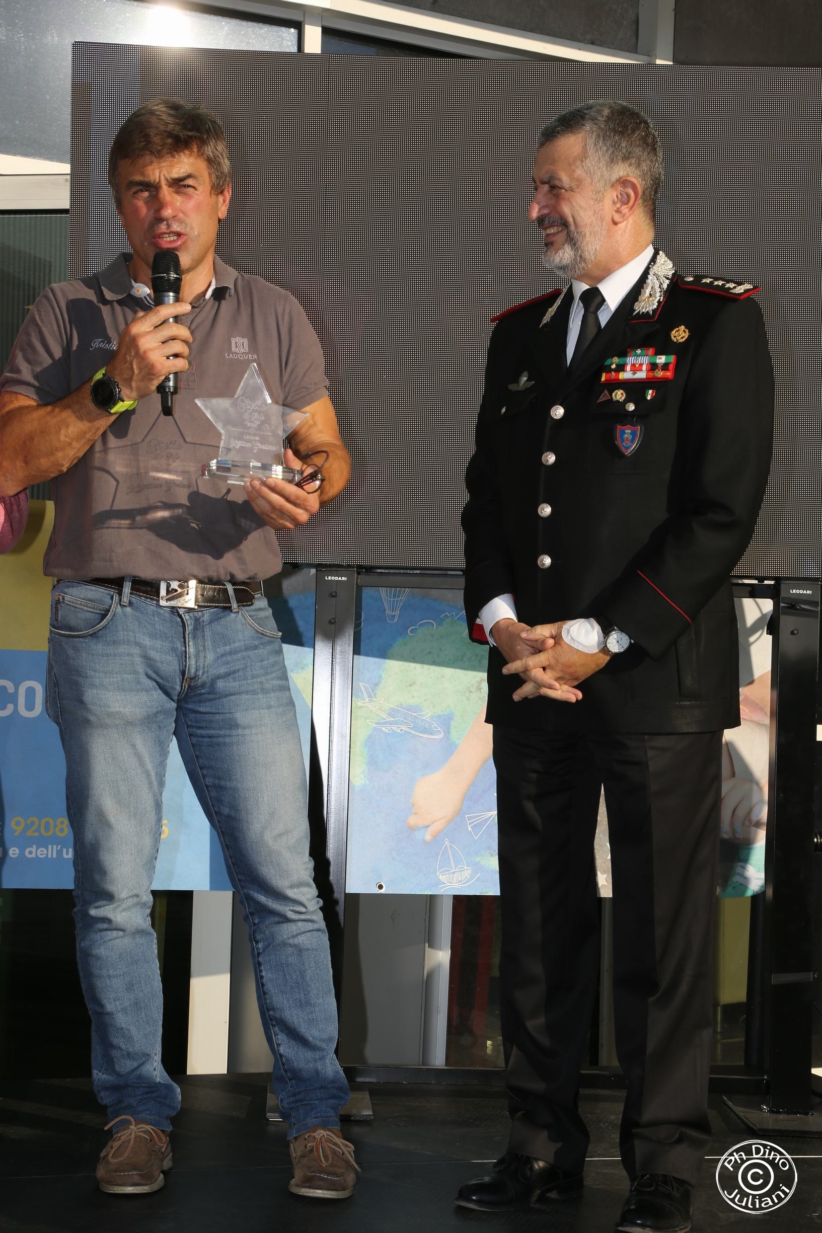 due persone sul palco, uno vestito in uniforme sorridente. L'altro tiene un premio a forma di stella e parla al microfono.