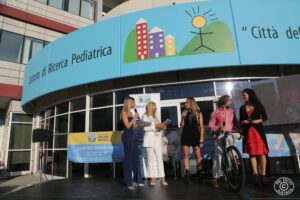 Quattro donne sono in piedi e parlano su un palco all'esterno di un istituto di ricerca pediatrica; un uomo tiene una bicicletta.