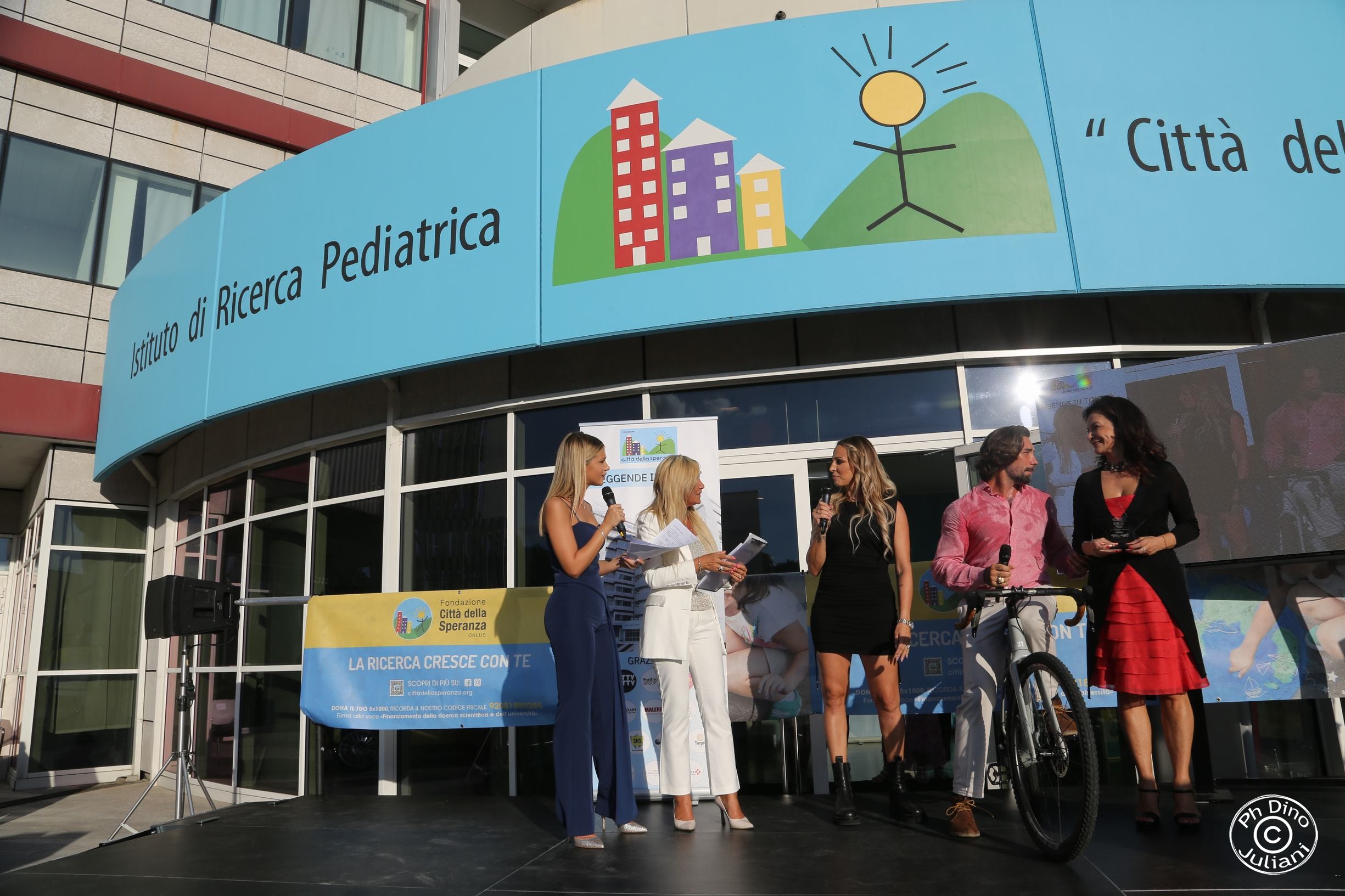 Quattro donne sono in piedi e parlano su un palco all'esterno di un istituto di ricerca pediatrica; un uomo tiene una bicicletta.