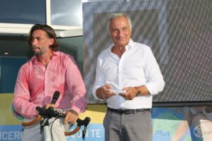 Premiazione all’evento “Leggende in Torre” della Città della Speranza, con consegna di un riconoscimento a un partecipante, affiancato da una persona su una bicicletta.