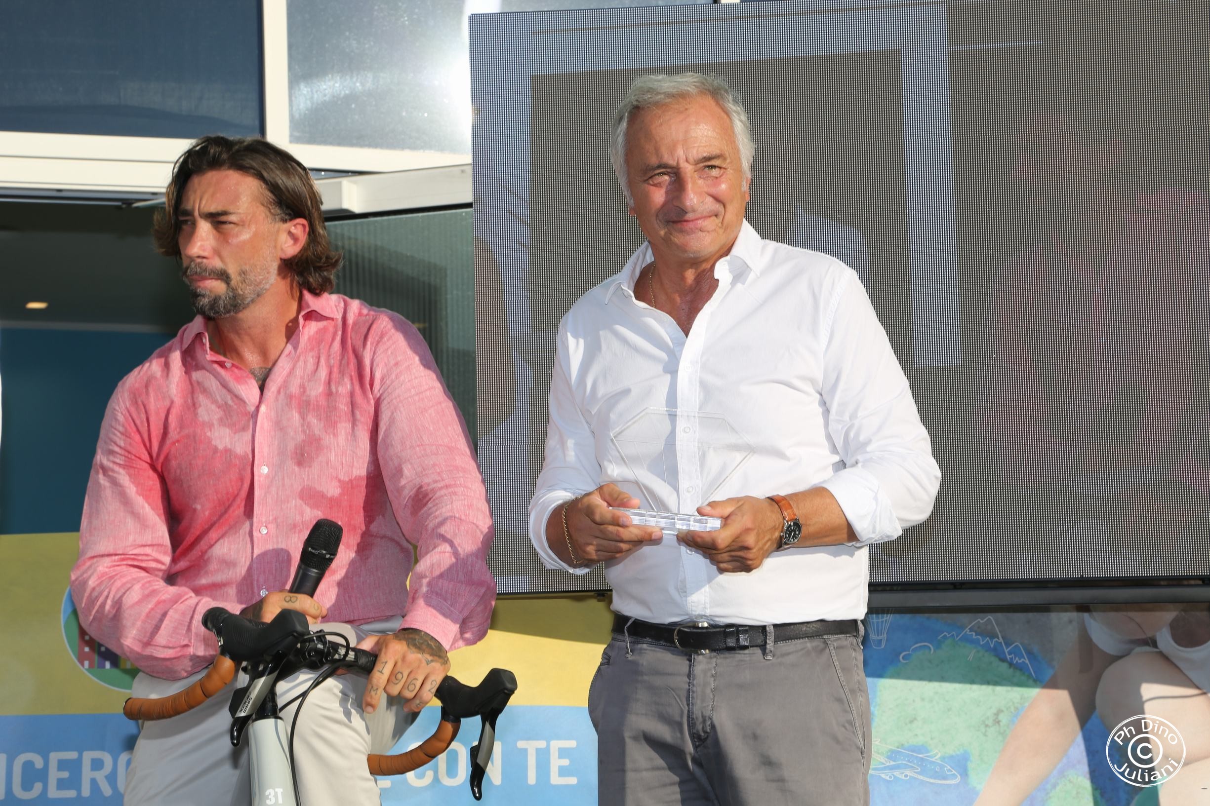 Premiazione all’evento “Leggende in Torre” della Città della Speranza, con consegna di un riconoscimento a un partecipante, affiancato da una persona su una bicicletta.