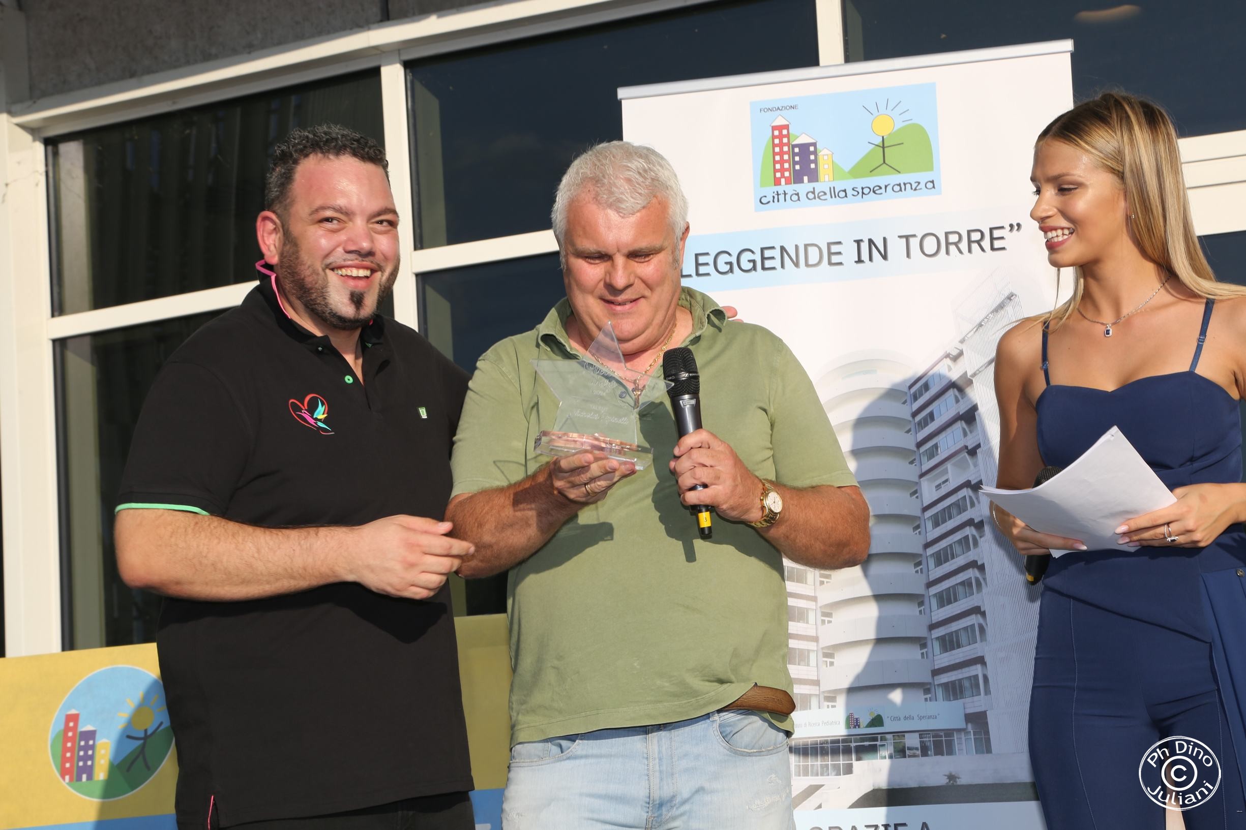 L’uomo al centro tiene in mano un trofeo trasparente e un microfono, affiancato da un uomo con polo nera e una donna in abito blu che tiene dei fogli.