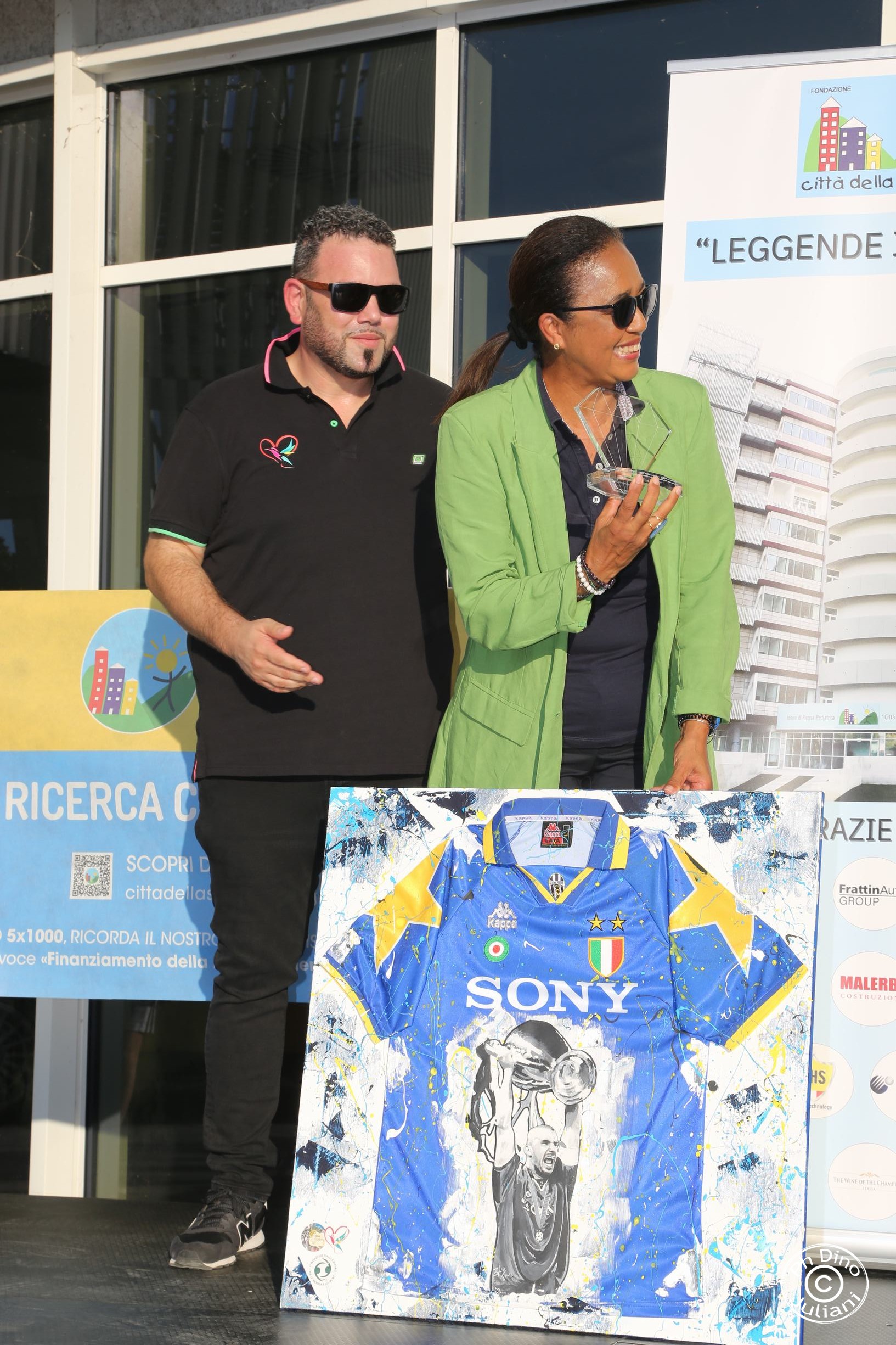 Due persone sul palco per l'evento leggende in torre presso Città della Speranza. Una donna tiene un oggetto in mano a forma di una stella Econ l'altra mano tiene un quadro con una maglietta di calcio