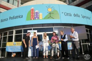 Un gruppo di persone è riunito davanti all'ingresso dell'Istituto di Ricerca Pediatrica "Città della Speranza". Uno regge una bicicletta, altri posano sorridendo per la foto. Sullo sfondo, il logo colorato dell’istituto è ben visibile sull’insegna azzurra sopra l’entrata.