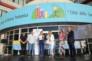 Evento all’aperto davanti all’Istituto di Ricerca Pediatrica "Città della Speranza". Un gruppo di persone è riunito su un palco. Due persone tengono un microfono. Una persona è sopra una bicicletta