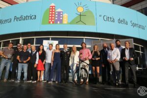 Una persona al centro tiene un microfono e parla al pubblico seduto su sedie bianche. Dietro il gruppo c’è una bicicletta esposta.