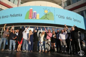 Palco davanti all'ingresso di Città della Speranza dove indicano con le dita l'insegna Città della Speranza