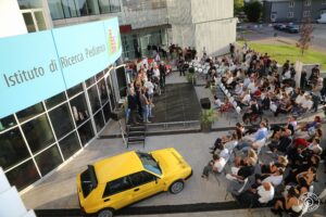 immagine dall'alto che si vedono le persone sul palco, il pubblico e una macchina gialla